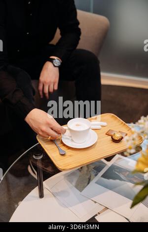 Man taking cappuccino from wooden tray with chocolates in lounge, glass table, close up Stock Photo