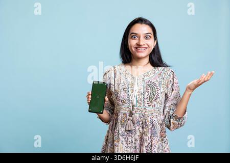 Happy indian woman presenting green screen on her smartphone, posing against the blue background. Joyous girl holding copy space device used for advertising brands in a commercial. Stock Photo