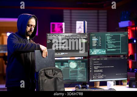 Hacker packing laptop after finishing coding stealth malware to evade security. Evil man putting notebook in backpack after finishing work on script hiding malicious code from detection systems Stock Photo