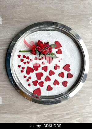 Round metal tray with powdered sugar & decorated with red heart icing sugar cutouts sugarcraft & small artificial flower arrangement valentines day Stock Photo