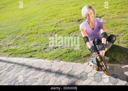 Senior woman sitting at park in lavender fixing skates with yellow wheels and pads, copy space. Inline, athletic, outdoor, mature, lifestyle, sunlight Stock Photo