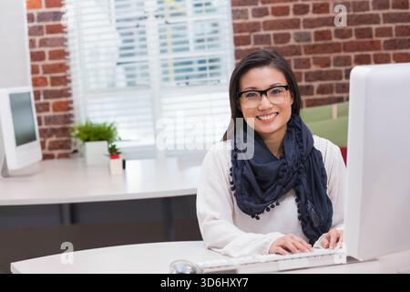 Mid-adult Asian woman wearing glasses and navy scarf typing at office desk with monitor keyboard. Professional, workspace, sunlit, modern, brickwall, Stock Photo