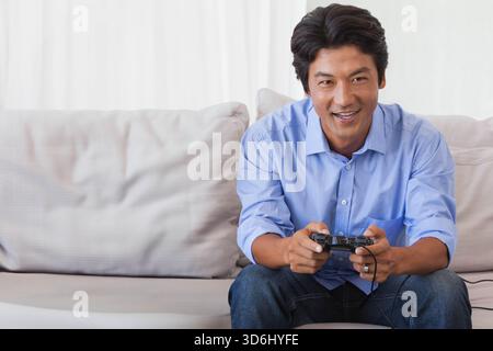 Mid adult Asian man wearing light blue shirt and dark jeans, playing wired controller on sofa. Gaming, relaxation, home, livingroom, ambience, minimal Stock Photo