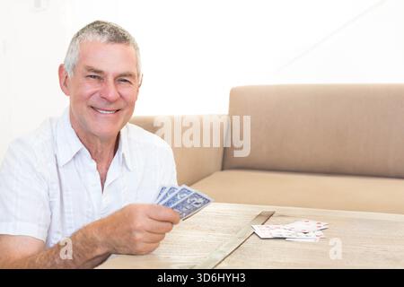 Senior man sitting at wooden table holding playing cards with beige sofa behind, copy space. Elderly, gentleman, retiree, lounge, home, minimal, calm Stock Photo