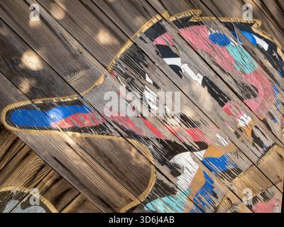 Close-up of a weathered, hand‑painted duck design on diagonal wooden planks. Layers of peeling paint in red, blue, teal, black, white, and gold. Stock Photo