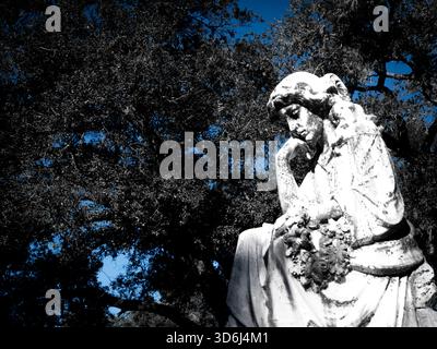 Weathered stone statue of a contemplative woman resting her head on one hand and holding a floral wreath. The figure is highlighted against dark. Stock Photo