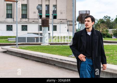 Man in coat and jeans walking through an urban park, thoughtful and looking toward the city skyline on a cloudy day Stock Photo
