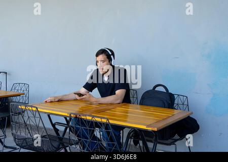 Young man in headphones relaxing at an outdoor cafe table, focused on his smartphone, enjoying music and modern urban downtime Stock Photo