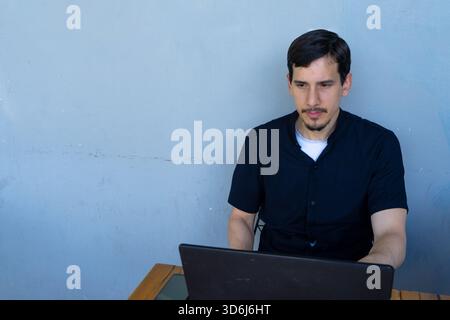 Man focused on laptop at a wooden desk, remote work or study in a casual cafe.home office setting, serious and contemplative user Stock Photo