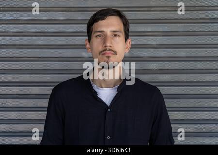 Young man with goatee and neutral, serious expression standing against a grey metal roller shutter, urban casual portrait, upper body Stock Photo