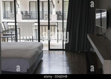 Calm hotel room interior showing comfortable bed, wood floor, and television. large glass window provides relaxing view of building and balcony outsid Stock Photo