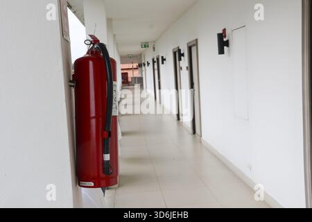 Red fire extinguisher for safety in long, quiet, empty building hallway. white corridor interior with tiled floor and row of doors creates lonely, pea Stock Photo