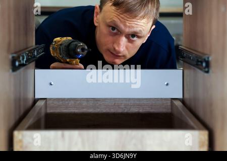 Close-up of carpenter drilling inside furniture drawer. Man looking into drawer while using power drill to install slides in wooden cabinet interior Stock Photo