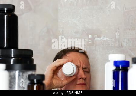 Man closely inspecting pill bottle label among supplements. Man squinting and holding white pill bottle close to eye, surrounded by various supplement Stock Photo