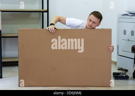 Man curiously peeking over large cardboard sheet in office. Young man in white t-shirt hiding and peeking over blank brown cardboard sheet indoors, co Stock Photo