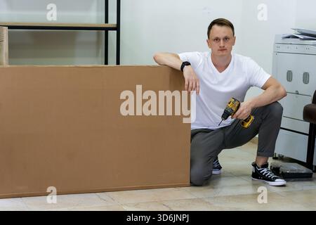 Young man crouching and holding drill next to large cardboard box in office setting. Casual man in white shirt kneels with power drill beside tall bro Stock Photo