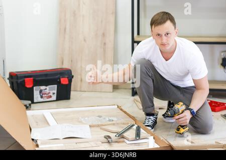 Young man giving thumbs up while assembling flat pack furniture with cordless drill. Man in casual clothes crouches with drill, giving thumbs up durin Stock Photo