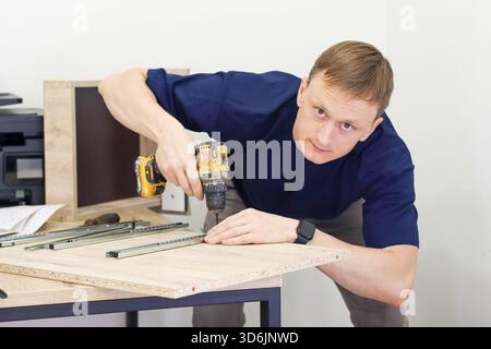 Man assembling flat pack furniture with cordless drill. Young man in casual clothes using cordless drill to assemble wooden cabinet indoors, DIY home Stock Photo