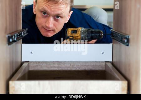 Carpenter installing drawer slide in wooden cabinet. Handyman using cordless drill to mount metal drawer slide inside modern wooden kitchen cabinet. Stock Photo
