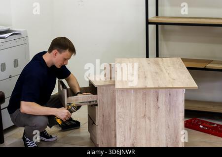 Man assembling wooden cabinet with cordless drill. Handyman in casual clothes using yellow cordless drill to install drawer slides on light wood cabin Stock Photo