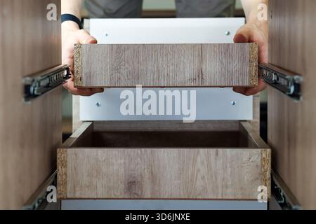 Person installing wooden drawer in cabinet. Close-up of hands installing a wooden drawer with metal slides into modern furniture cabinet Stock Photo