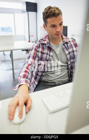 Man in 20s sitting in workspace using computer with keyboard and wired mouse, wearing plaid shirt. Adult, workstation, modern, bright, minimalist, off Stock Photo