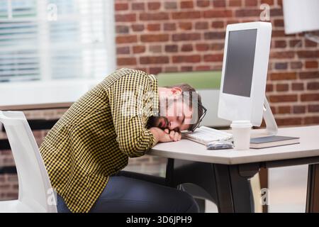 Adult African American man resting head on desk near monitor in office wearing checkered shirt. Coworking, brick, daylight, window, industrial, workst Stock Photo