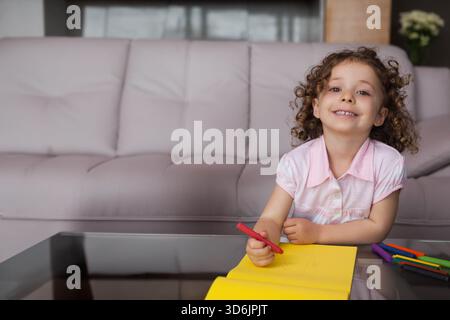 Female child drawing red marker on yellow pad at glass-top table in living room, copy space. Kid, curly, sofa, tufted, cozy, flowers, reflection Stock Photo