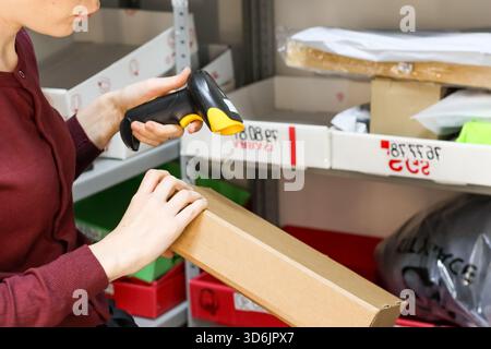 Female worker using handheld scanner on box label while holding package in storage rack Stock Photo
