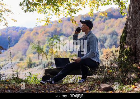A relaxed man in casual attire sits on wood log, drinks from coffee cup, views phone, laptop open nearby under autumn trees and hills Stock Photo