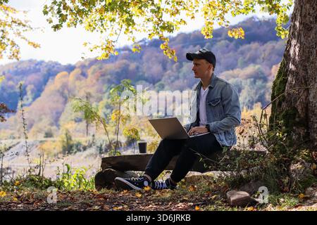Man in black cap and denim jacket uses laptop on knee while lounging on bench in sunny autumn forest, steam from coffee cup, backdrop of fiery orange Stock Photo