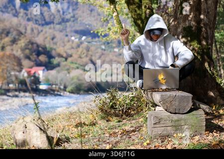 A masked man in hoodie sits by tree, right hand pointing upward, left on wrist, laptop on stump against autumn mountain and river scene. Stock Photo