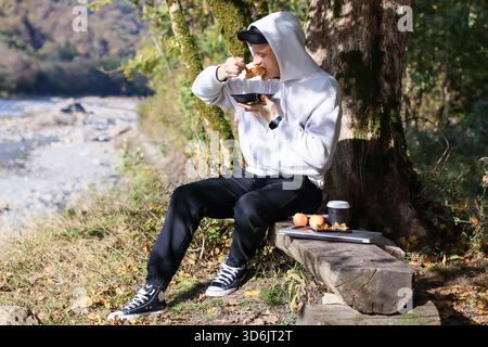 Man relaxing outdoors, consuming food from bowl on wooden seat by stream, with tech and snacks nearby. Stock Photo