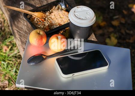 Top view of black food container with grain meal, wooden utensils, red apples, black coffee cup, black spoon, white phone on silver laptop on wooden l Stock Photo