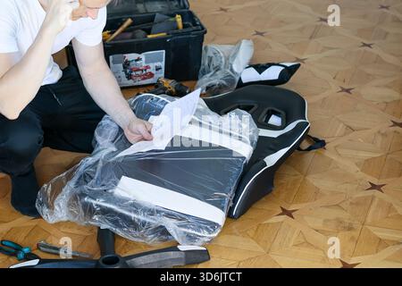 A man in casual clothes kneels on wooden floor, puzzled expression while studying blueprint for assembling ergonomic gaming chair with tools and packa Stock Photo