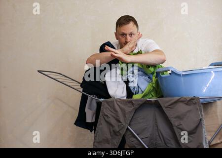 A young man makes a shushing gesture while surrounded by colorful clothes on a drying rack and a blue laundry basket in a home interior. (128 chars) Stock Photo