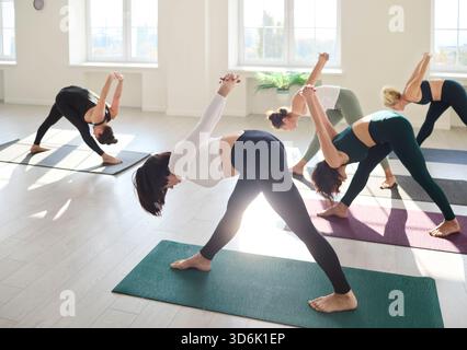 Group yoga class practicing wide-legged forward bends in bright sunny studio Stock Photo