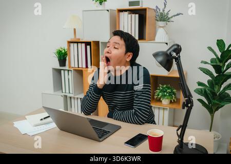 sleepy young man yawning while working with laptop computer at home office Stock Photo