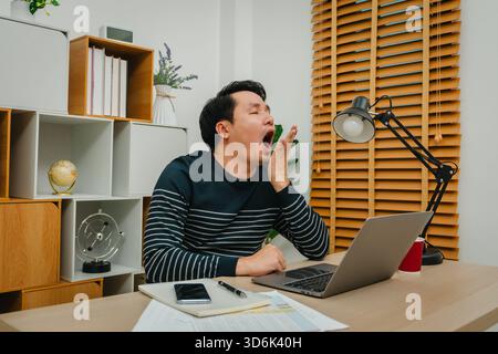 sleepy young man yawning while working with laptop computer at home office Stock Photo
