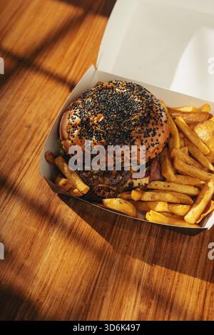 Delicious burger with black sesame bun and crispy fries served in a white takeout box on a sunlight wooden surface, photographed top view. Stock Photo