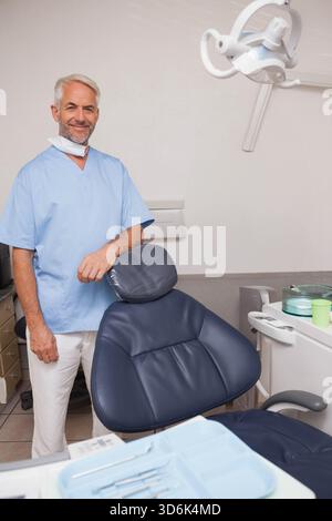 Senior male dentist standing by navy-blue chair in treatment room, wearing scrubs, showing tools. Dentistry, clinic, professional, modern, minimalist, Stock Photo