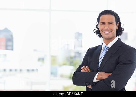 Man standing with arms crossed wearing pinstripe suit, tie in office by grid window, copy space. Headshot, professional, confident, urban, modern, min Stock Photo