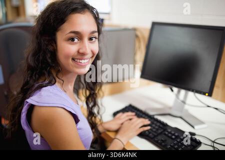 Female teen sitting at lab typing on keyboard, monitor and mouse, wearing lavender top, copy space. Woman, workstation, desk, classroom, education, te Stock Photo