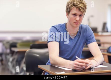 Male student in his twenties sitting at classroom desk holding pen over paper, wearing blue V-neck. Learner, adult, concentration, study, education, p Stock Photo