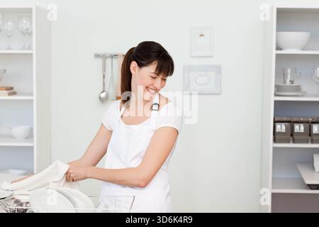 Woman wearing white apron drying plates with white dish towel in bright kitchen near dish rack. Minimalist, organized, welllit, glassware, shelving, c Stock Photo