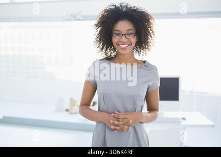 African American woman standing centered in office smiling clasping hands by desk with glasses. Workspace, professional, modern, bright, minimalist, g Stock Photo
