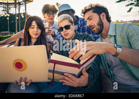 Diverse group of friends work together using a laptop and books outside in a park, sharing ideas and collaborating on a project. Stock Photo