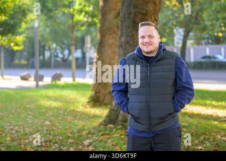 Relaxed young man standing outdoors in a sunny park with hands in pockets, surrounded by green trees and autumn leaves. Stock Photo