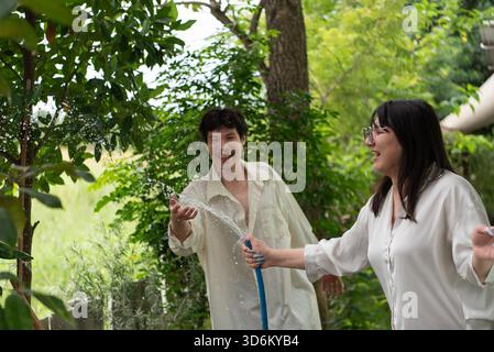 Two LGBTQ friends, one with long hair and the other with short hair, enjoy watering the garden together. They share a fun moment outdoors, taking care Stock Photo