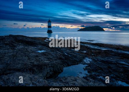Morning light over the lighthouse at Penmon Point with seaweed-covered rocks in the foreground and Puffin Island beyond under a layered sky Stock Photo
